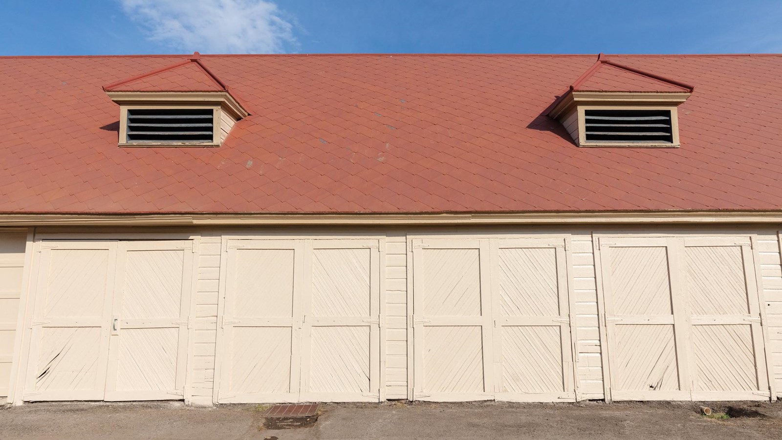 A historic building with pale yellow siding, four large barn doors, and a red shingle roof.