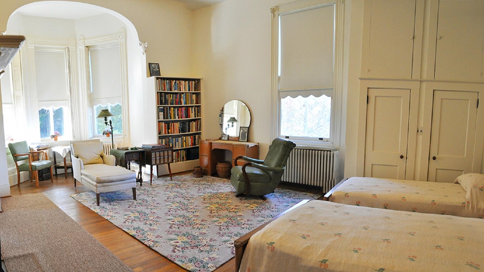 A large open room with two twin beds, a chaise lounge, vanity, bookshelves, and a alcove bay window.