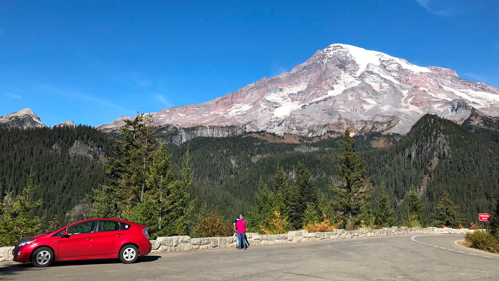 Two people look at a view of Mount Rainier from a paved viewpoint bordered by a rock wall.