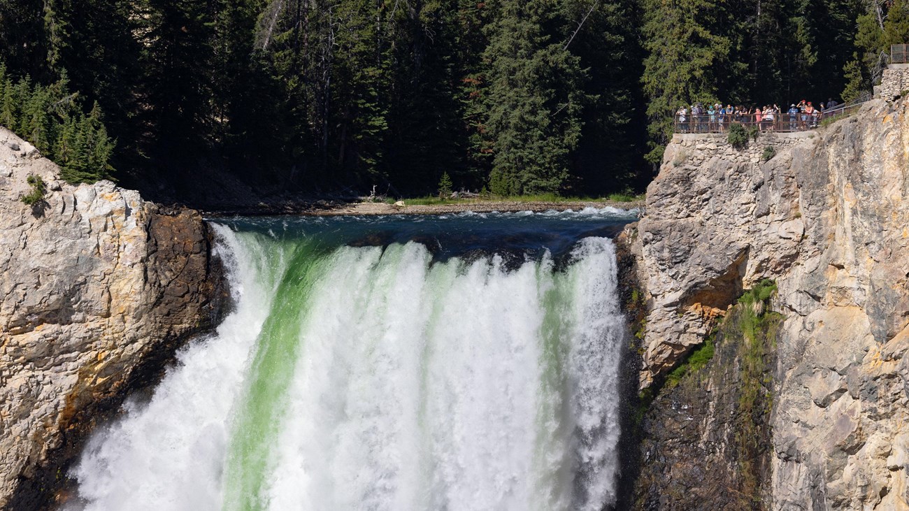 A group of visitors stand on a rock platform overlooking the top of a large waterfall.