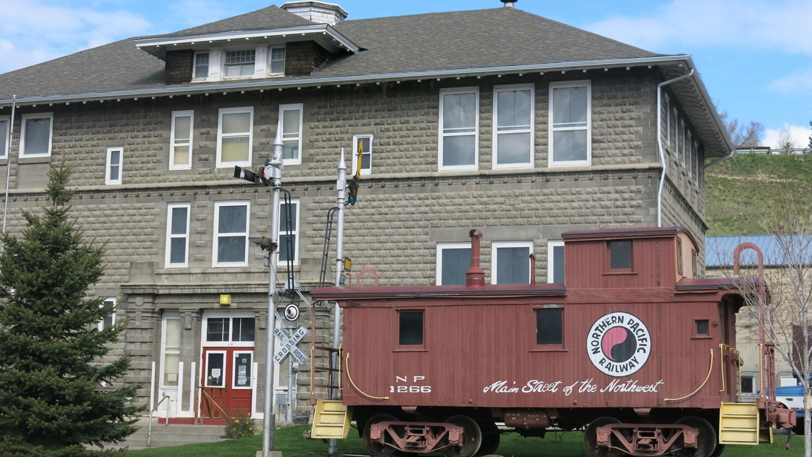 A three-story grey brick building with a red train caboose and a railway crossing sign in front..