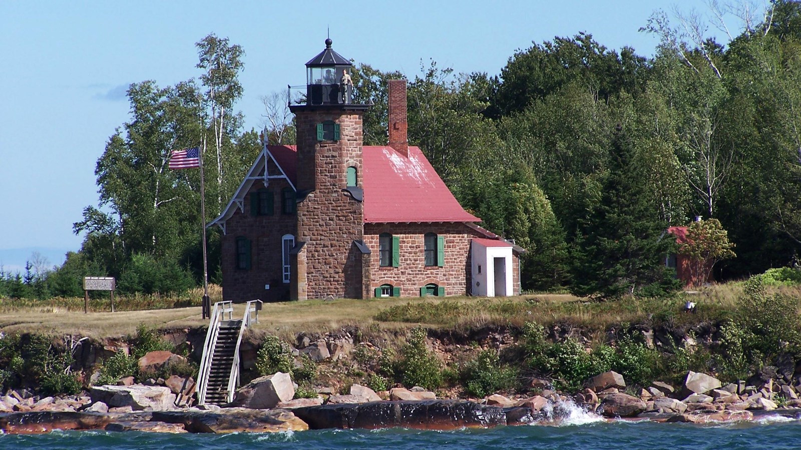 A brown stone lighthouse with a red roof on the shore of a lake surrounded by forest. 