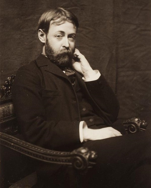 Black and white photograph of a young man. He is wearing dark clothes, seated, with hand near face.