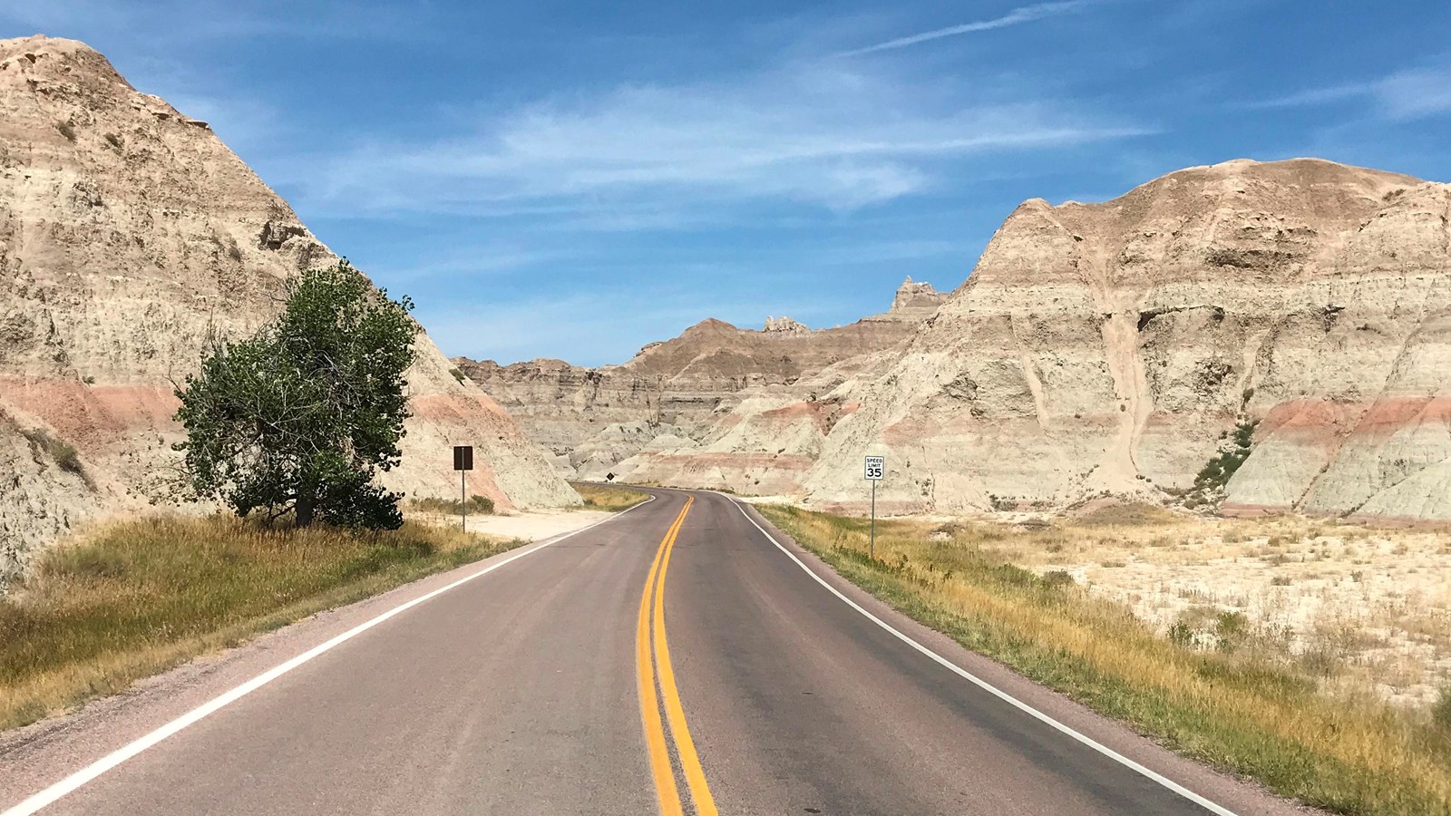 A paved two-lane highway traversed between two badlands buttes and sporadic cottonwoods along the ro