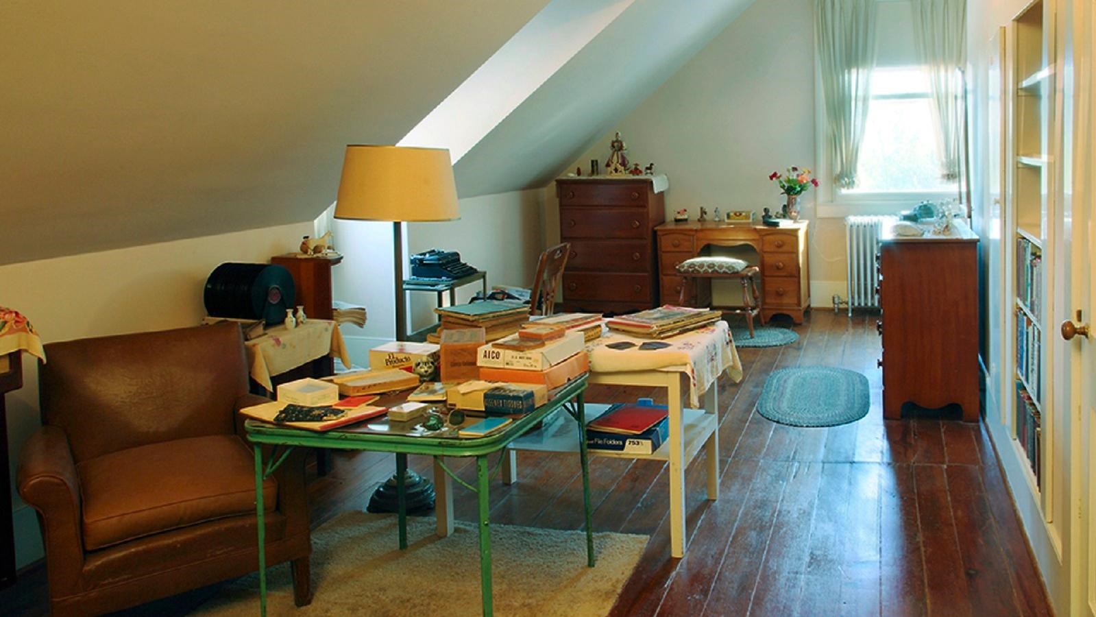 A room with a twin bed, books, chair, typewriter and card table, with knic knacs and personal items