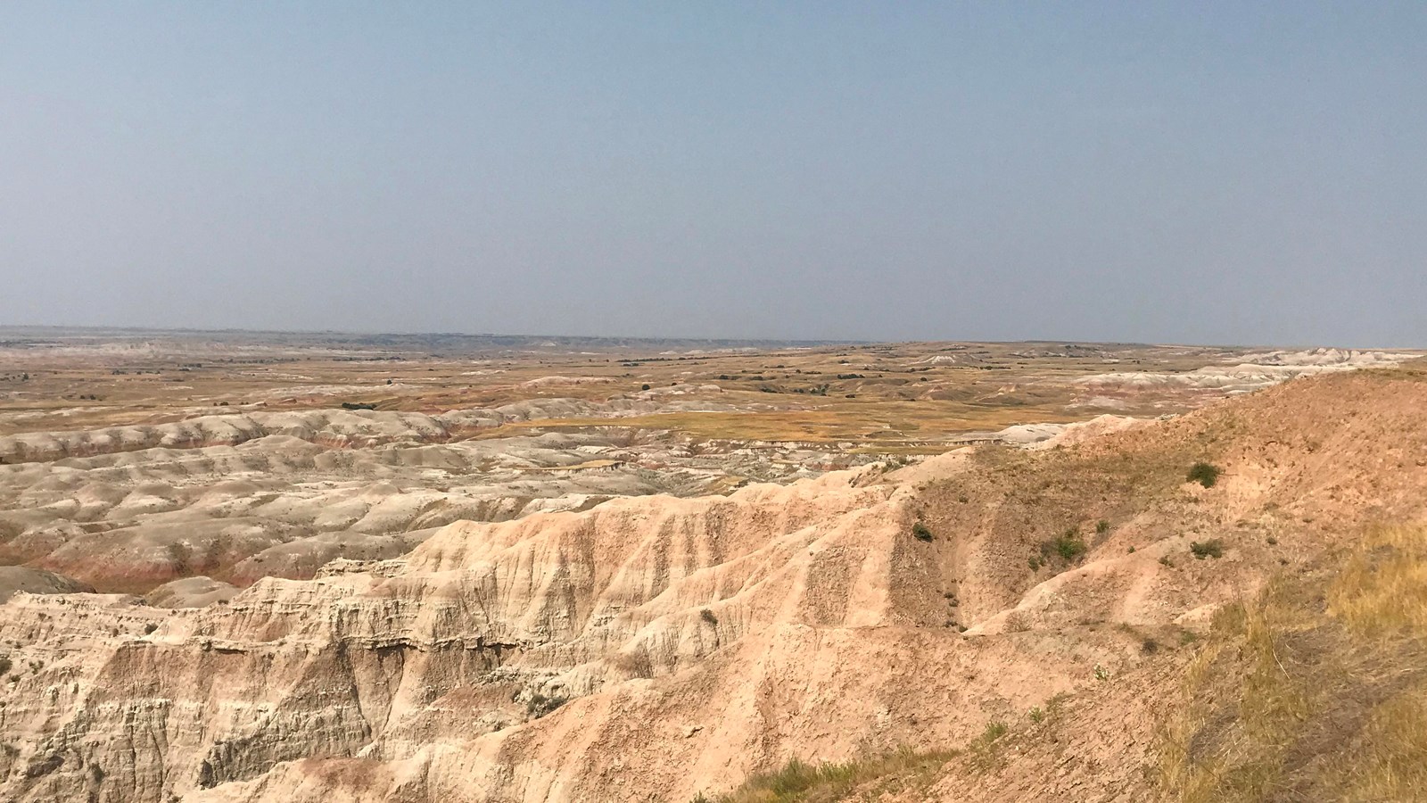 A vast expanse of badlands abuts against a wall with prairie grasses atop.