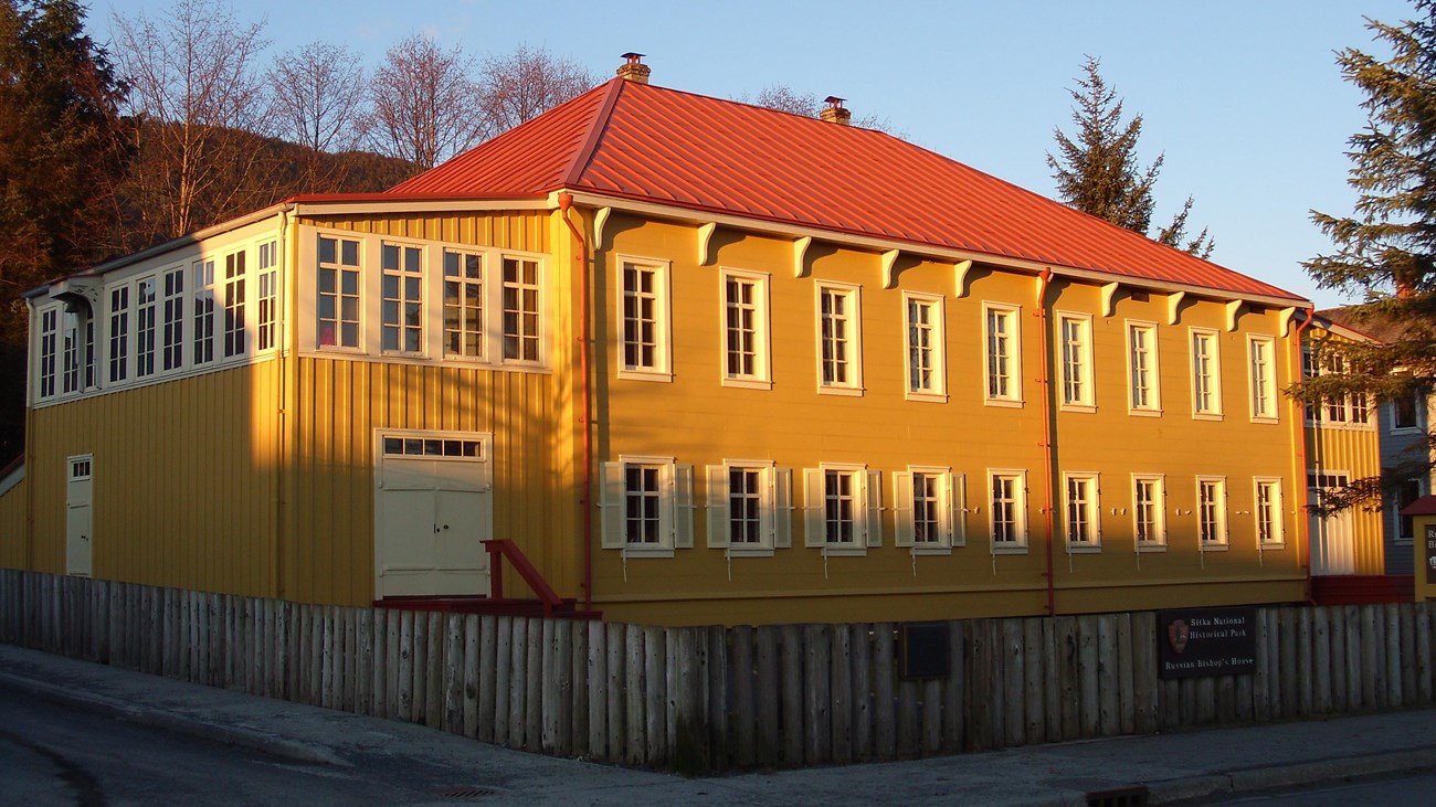 A two story yellow building with a red roof. A fence surrounds the building.