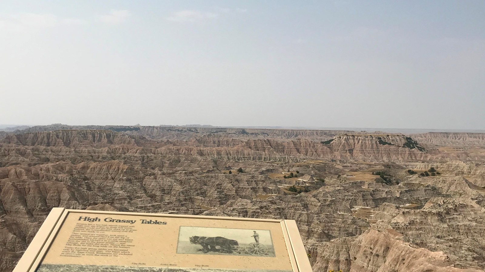 Wayside exhibit sign in foreground gives way to vast badlands wilderness under blue sky.