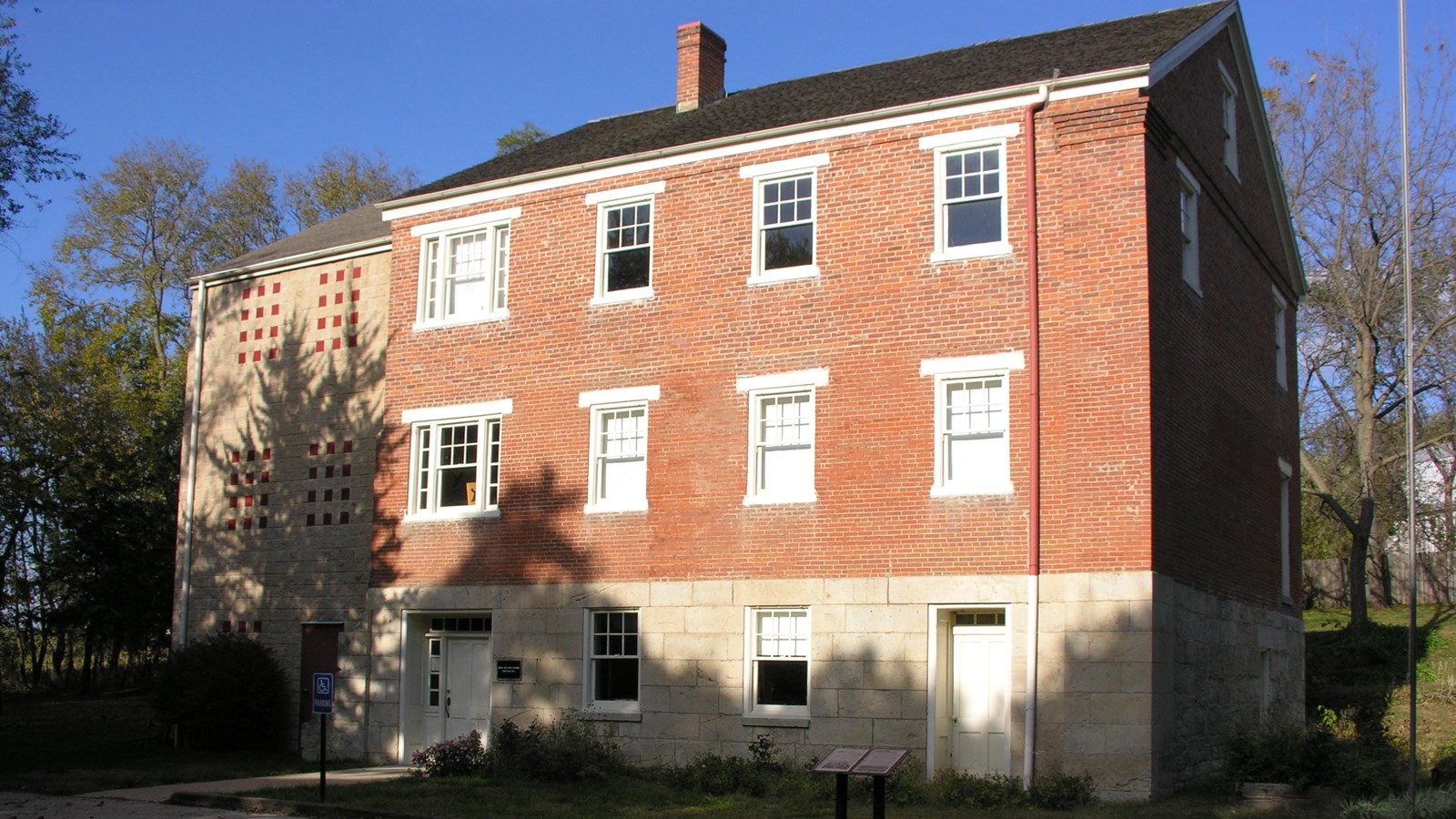 A 3-story brick and limestone building with a limestone addition stands in front of a parking area. 