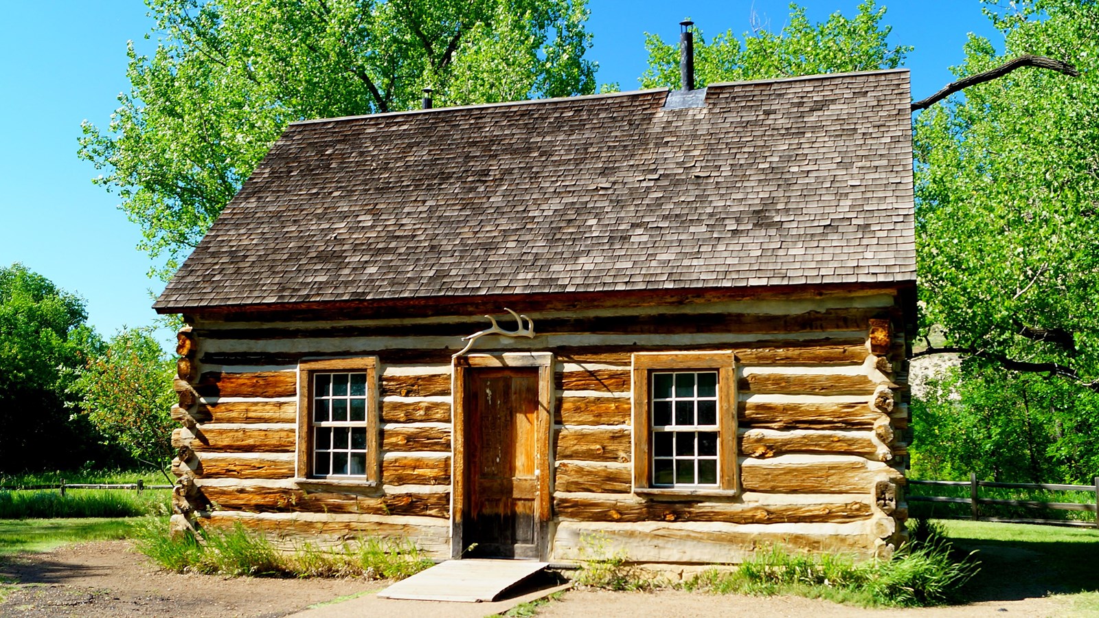 A log cabin sits in a grassy area. Trees and a fence are visible behind it, and a sidewalk in front.