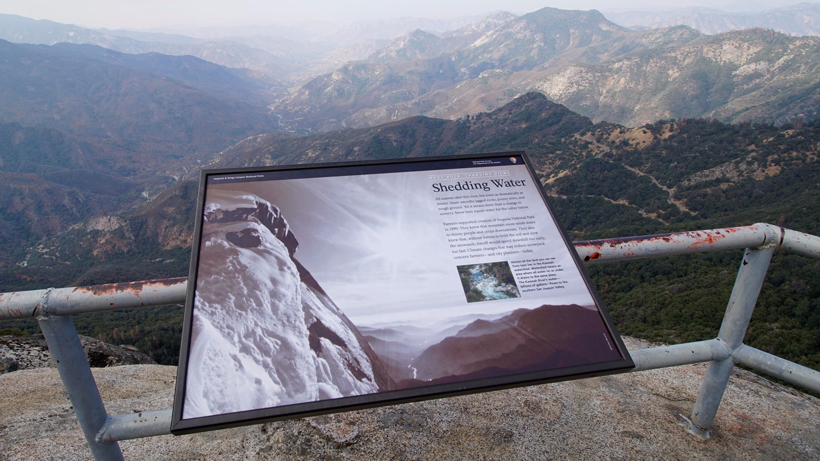 A metal panel sits on a metal railing high above the ground. In the distance mountains are visible