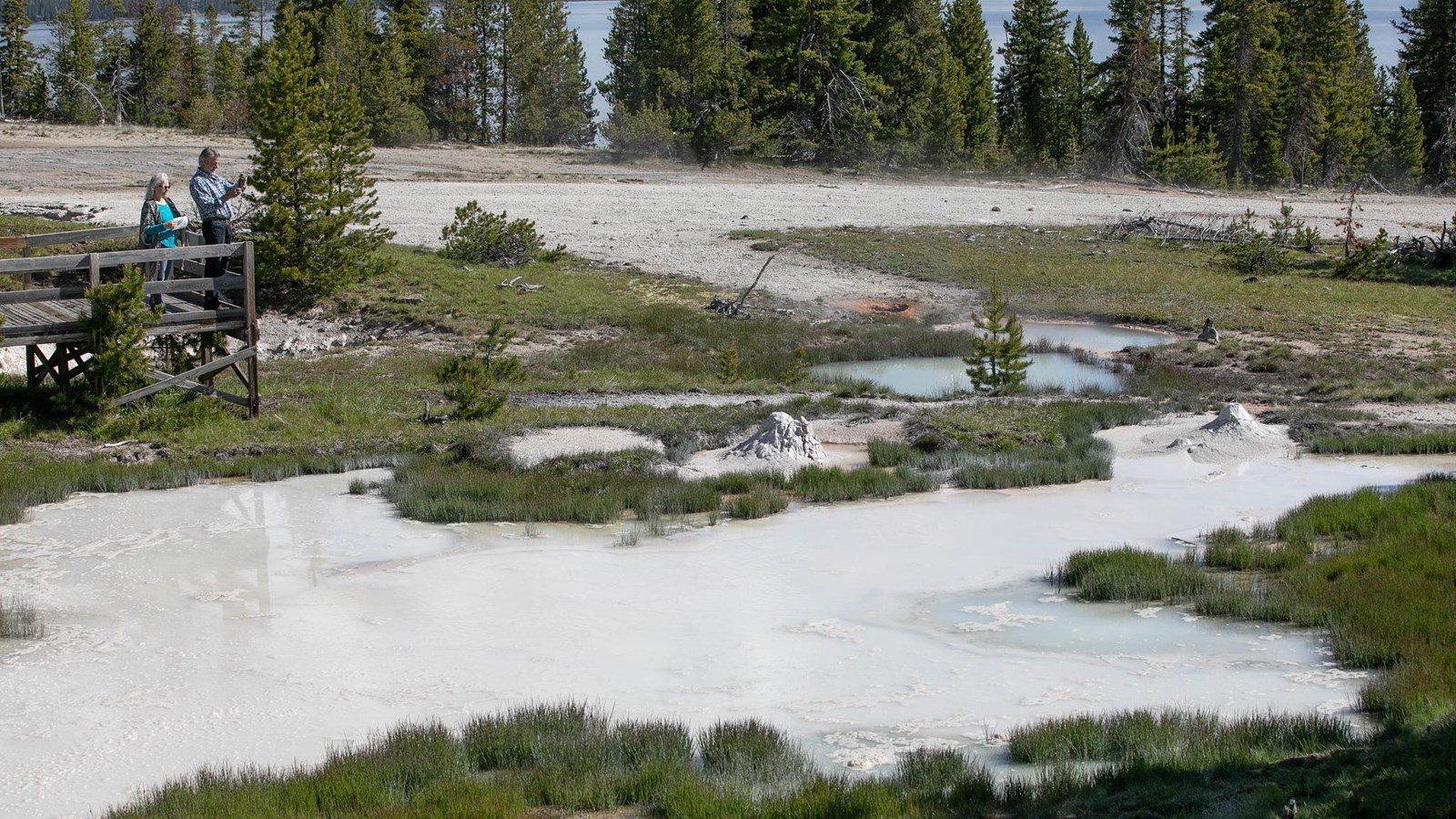 Two people stand on a boardwalk overlooking murky water with mounds or mud in a thermal area.