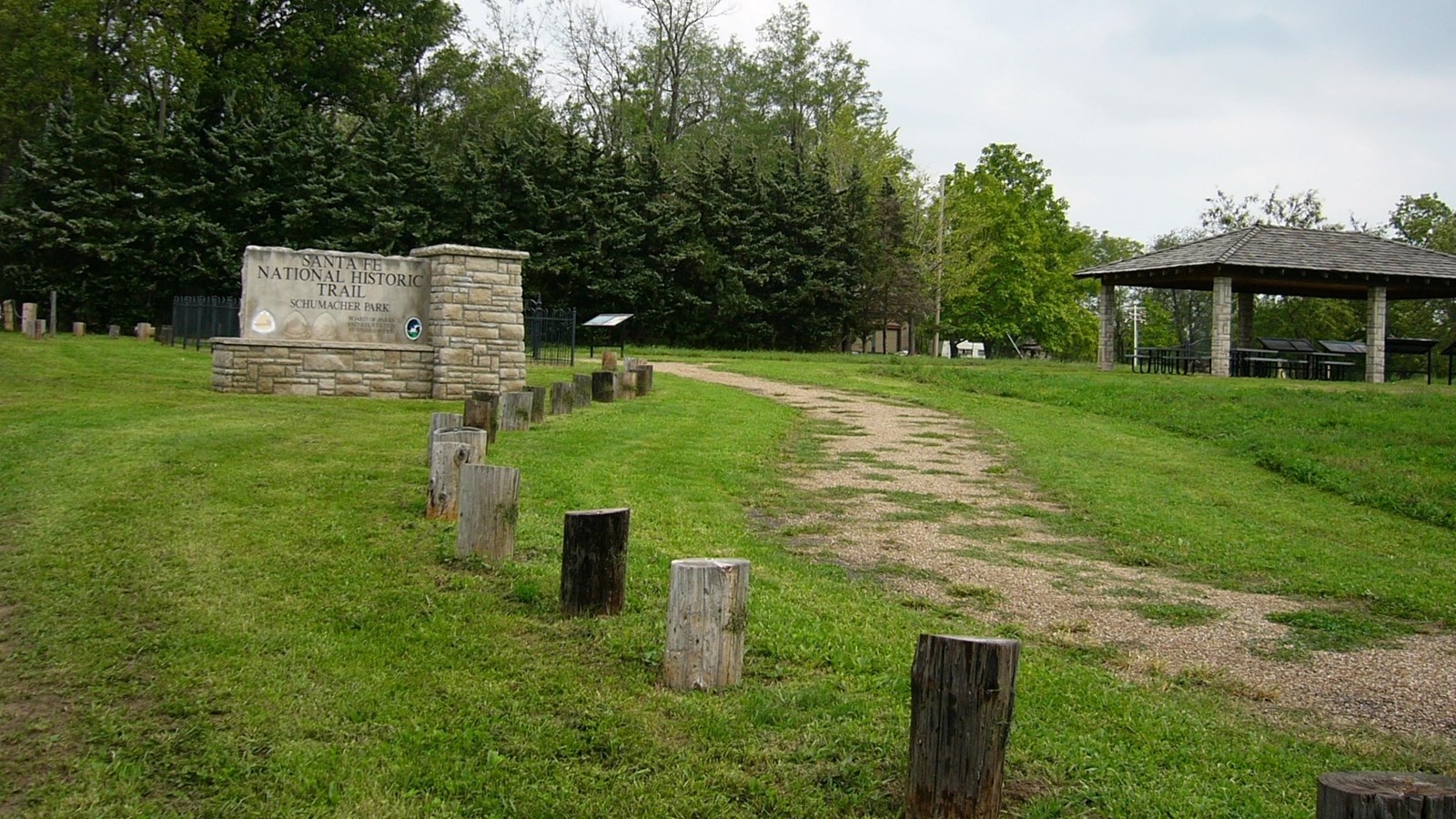 A path leads up a grassy hill, by a large stone sign, into a forested area.