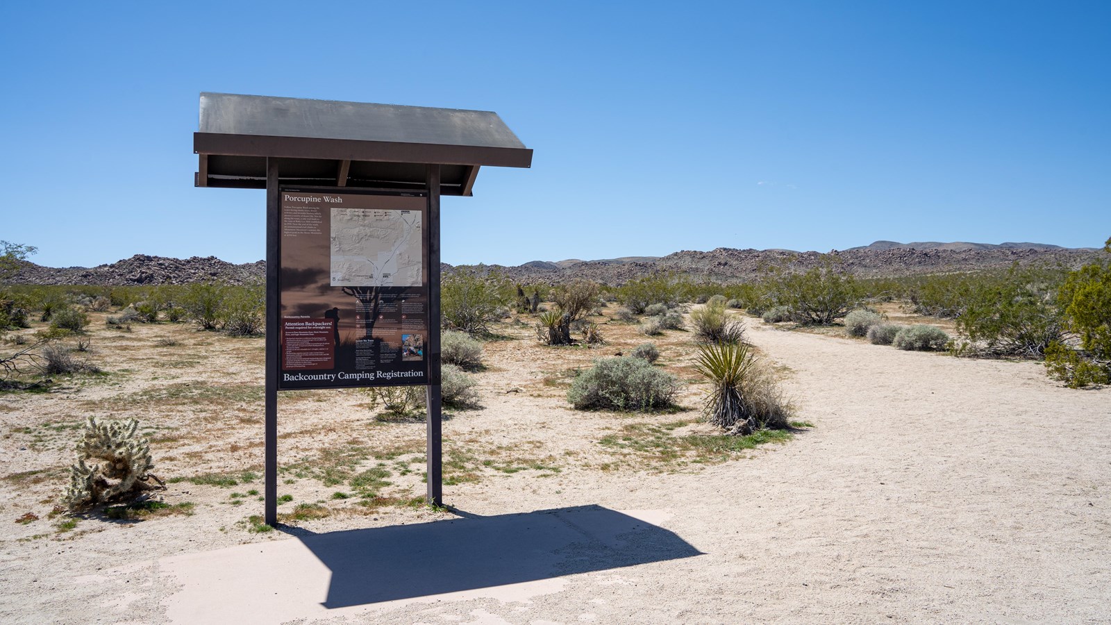 A wayside sign to the left of a trail leading into a desert landscape. Rocky hills in the distance.