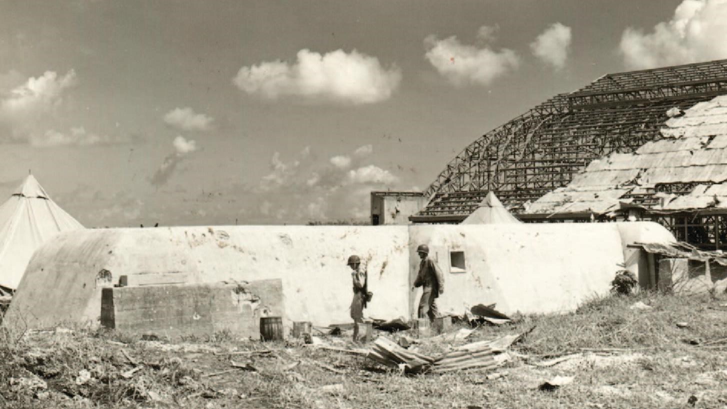Two US servicemen walk beside a Japanese air raid shelter. Behind is a war destroyed airplane hangar