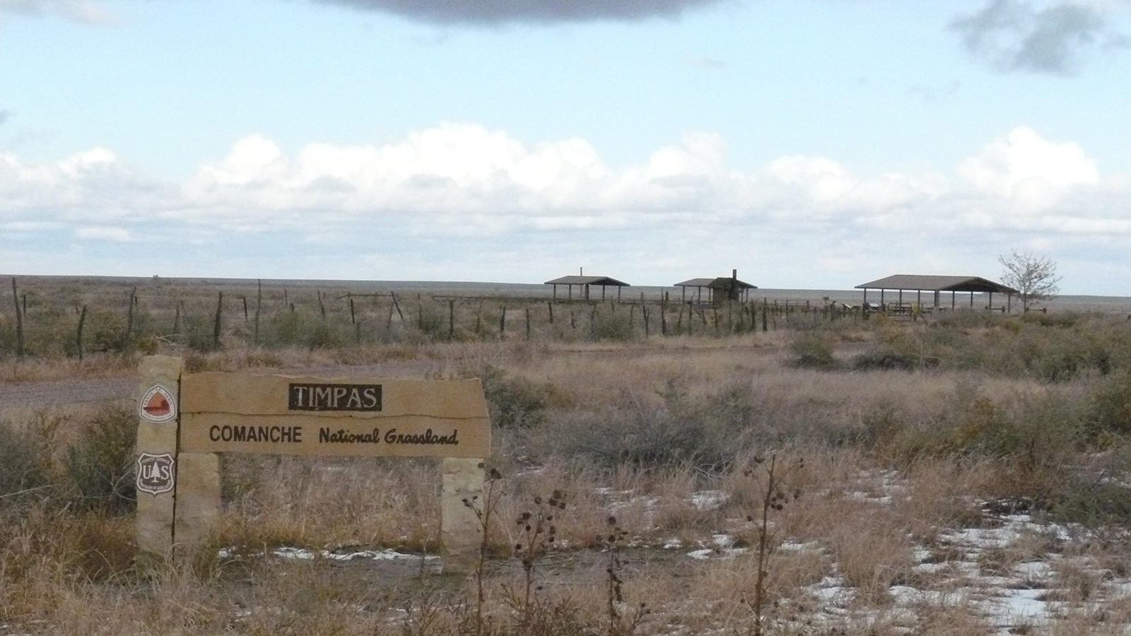 A sign shows the entrance to a distant picnic shelter on a flat prairie.