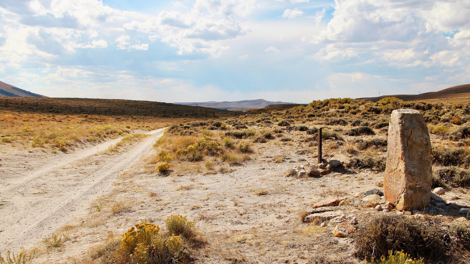 Yellow shrubs, a 2-track road, two stone monuments, and distant hills under a partly cloudy sky. 