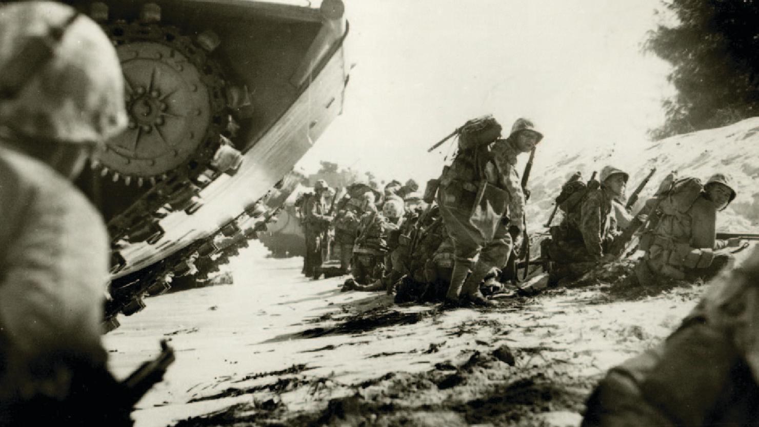 Many of marines in helmets, holding weapons crouch behind a sand barrier as they arrive on the beach