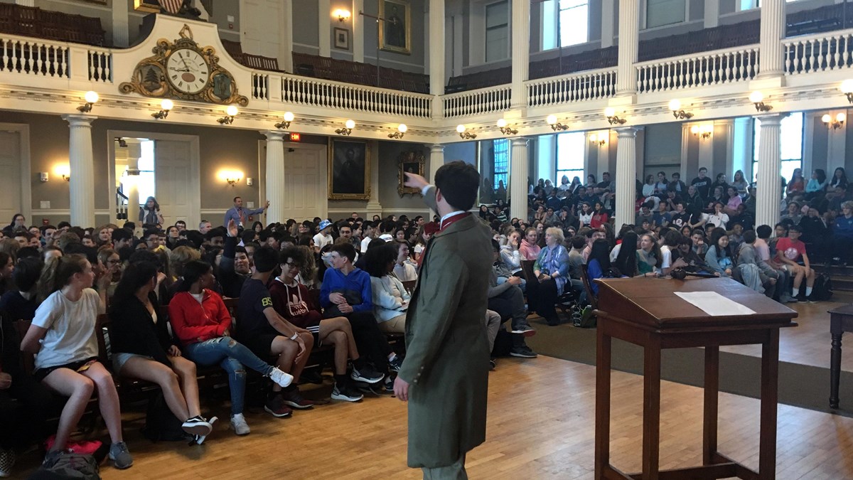 Interior of a large hall with a man speaking to a sitting audience of children.