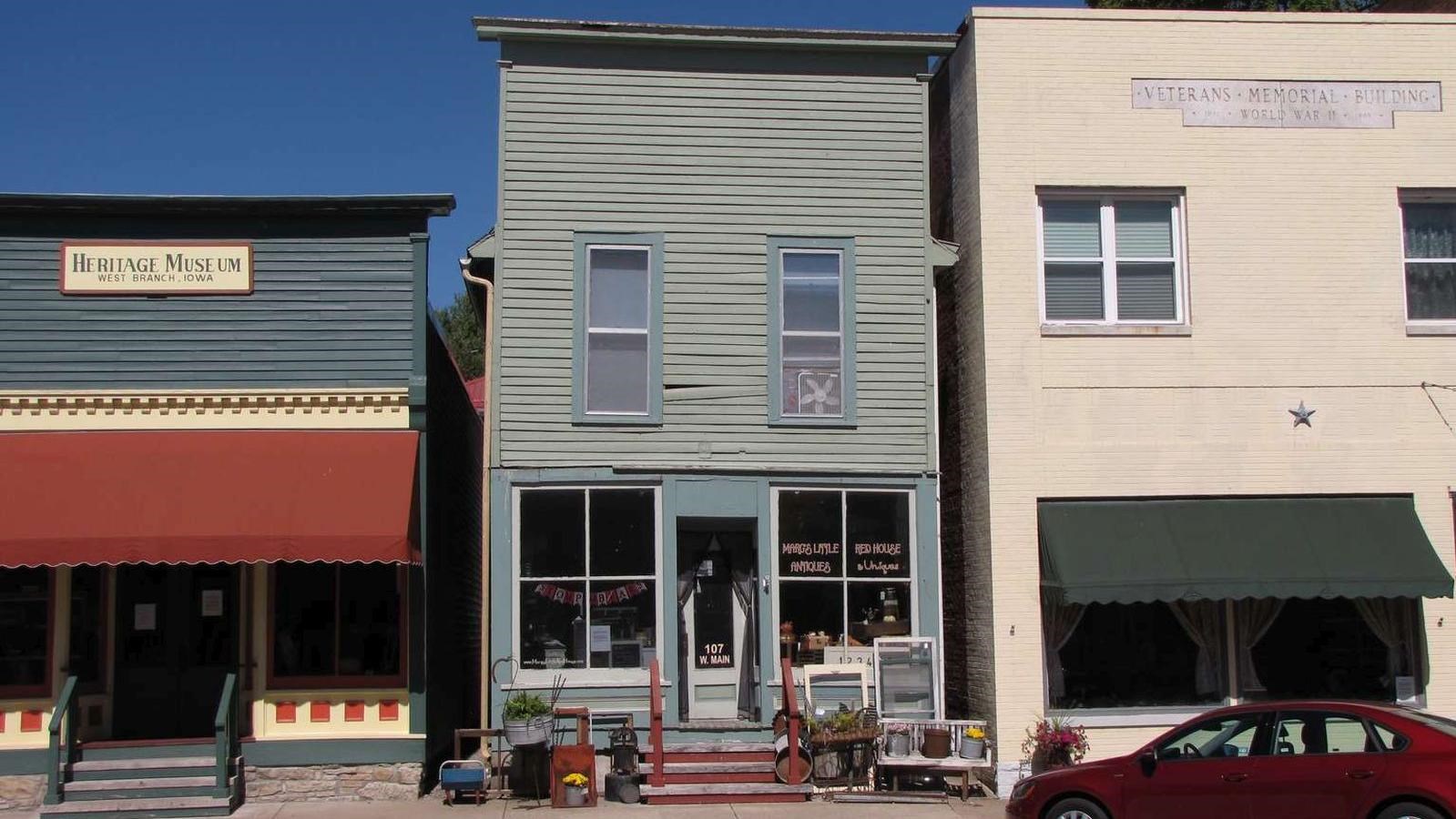 A narrow, two story light green building has wood siding and two windows above its storefront.