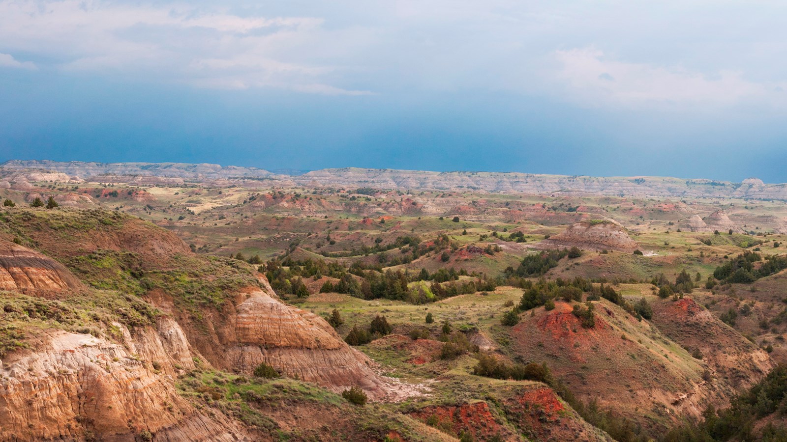 A view of colorful buttes under an overcast sky, with sunlight just breaking through