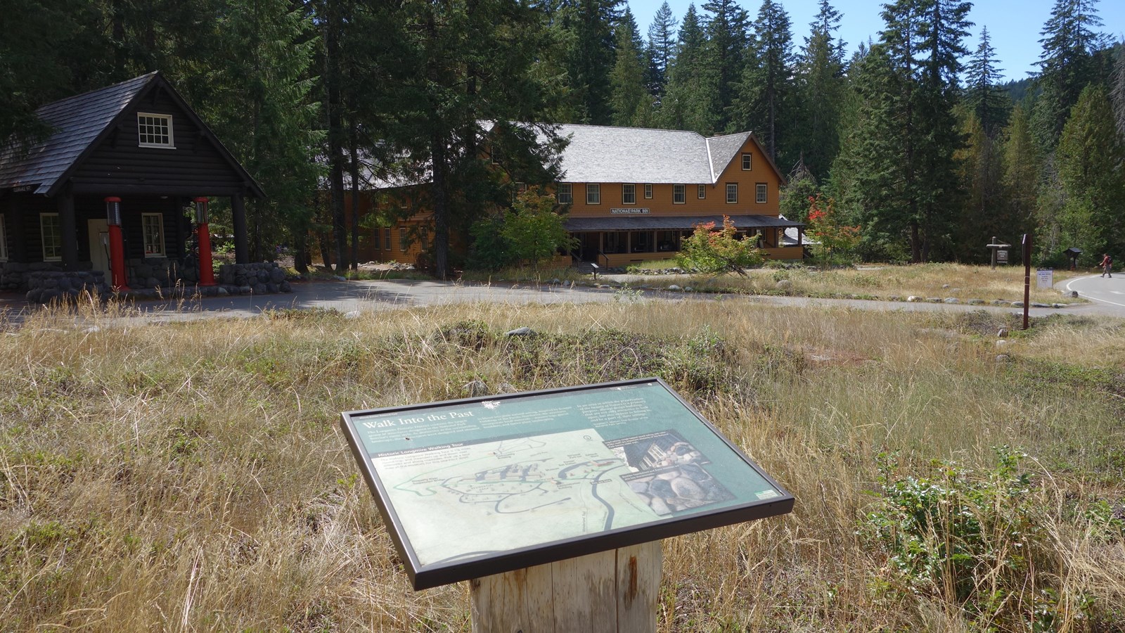 A wayside panel attached to a log stand in front of several historic buildings.