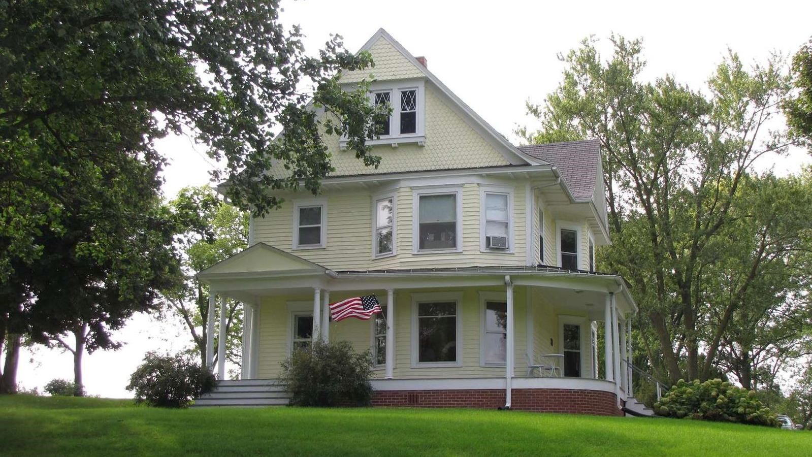 A large yellow house on a hill has three stories and a wraparound porch.