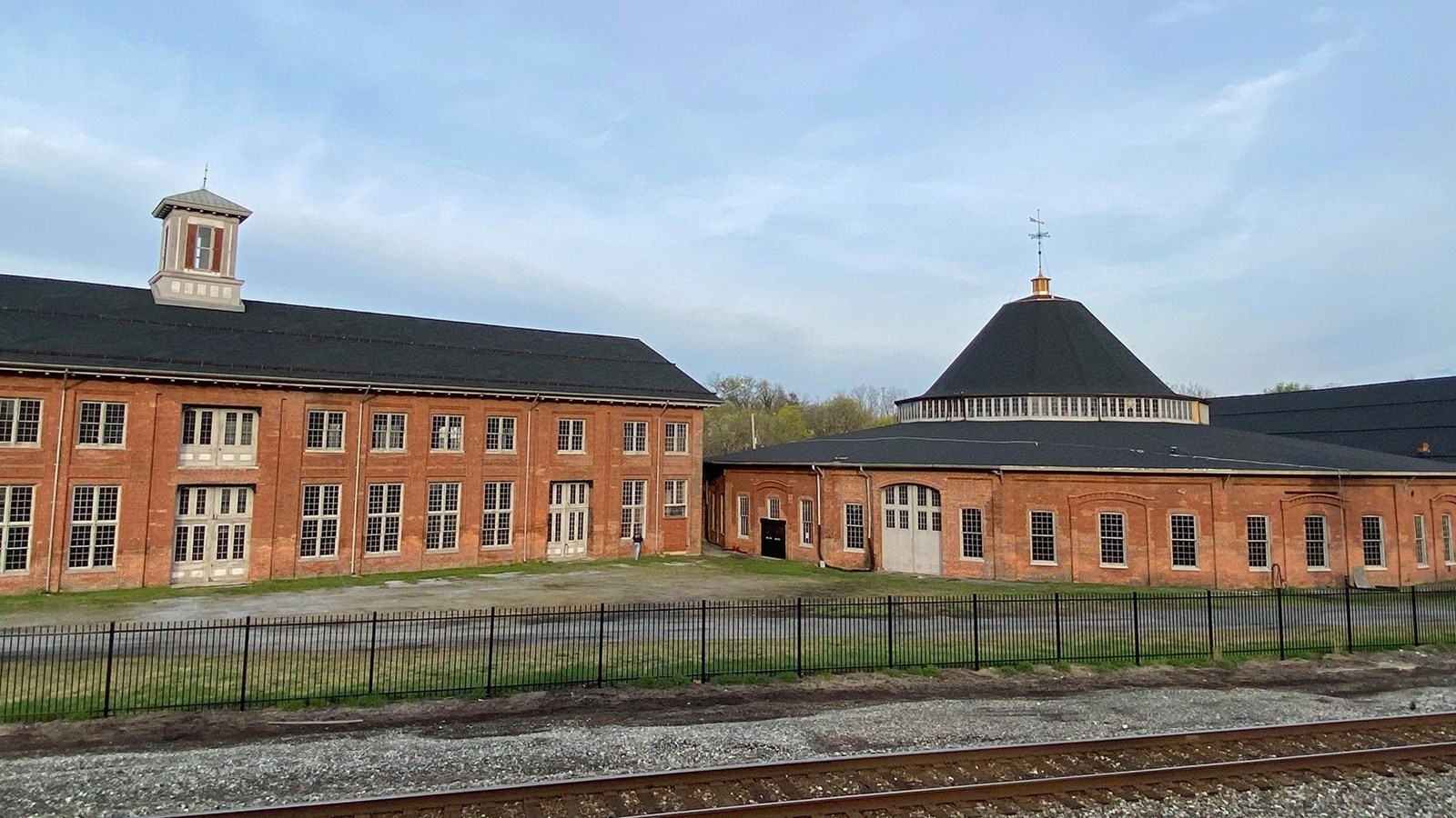 A sprawling brick building with rectangular windows set behind railroad tracks.