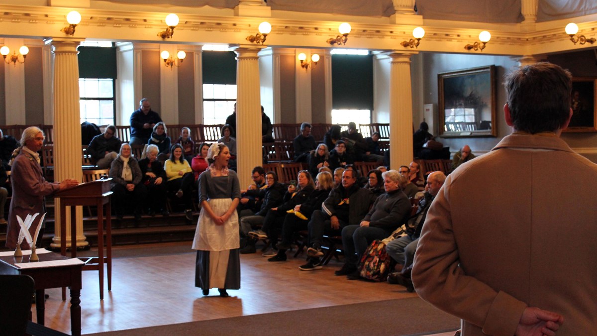 A large hall with a woman, wearing colonial period clothing, speaking to the seated audience.