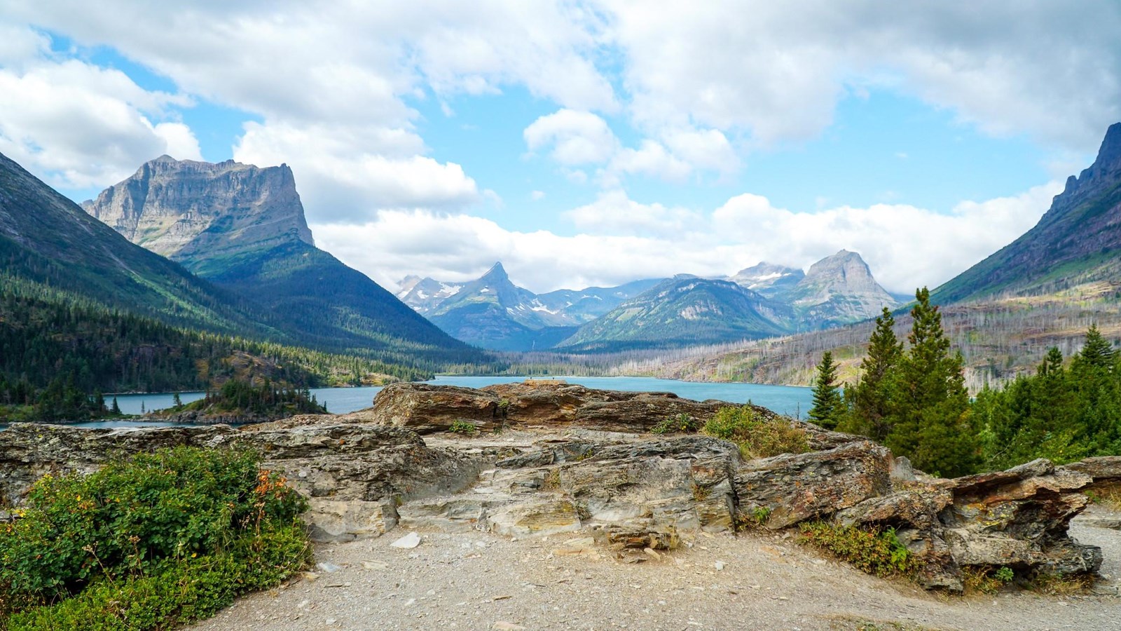 A view of distant mountains, a blue lake, and a rocky overlook.