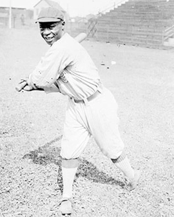 African American man in baseball uniform swinging a baseball bat facing toward the viewer.