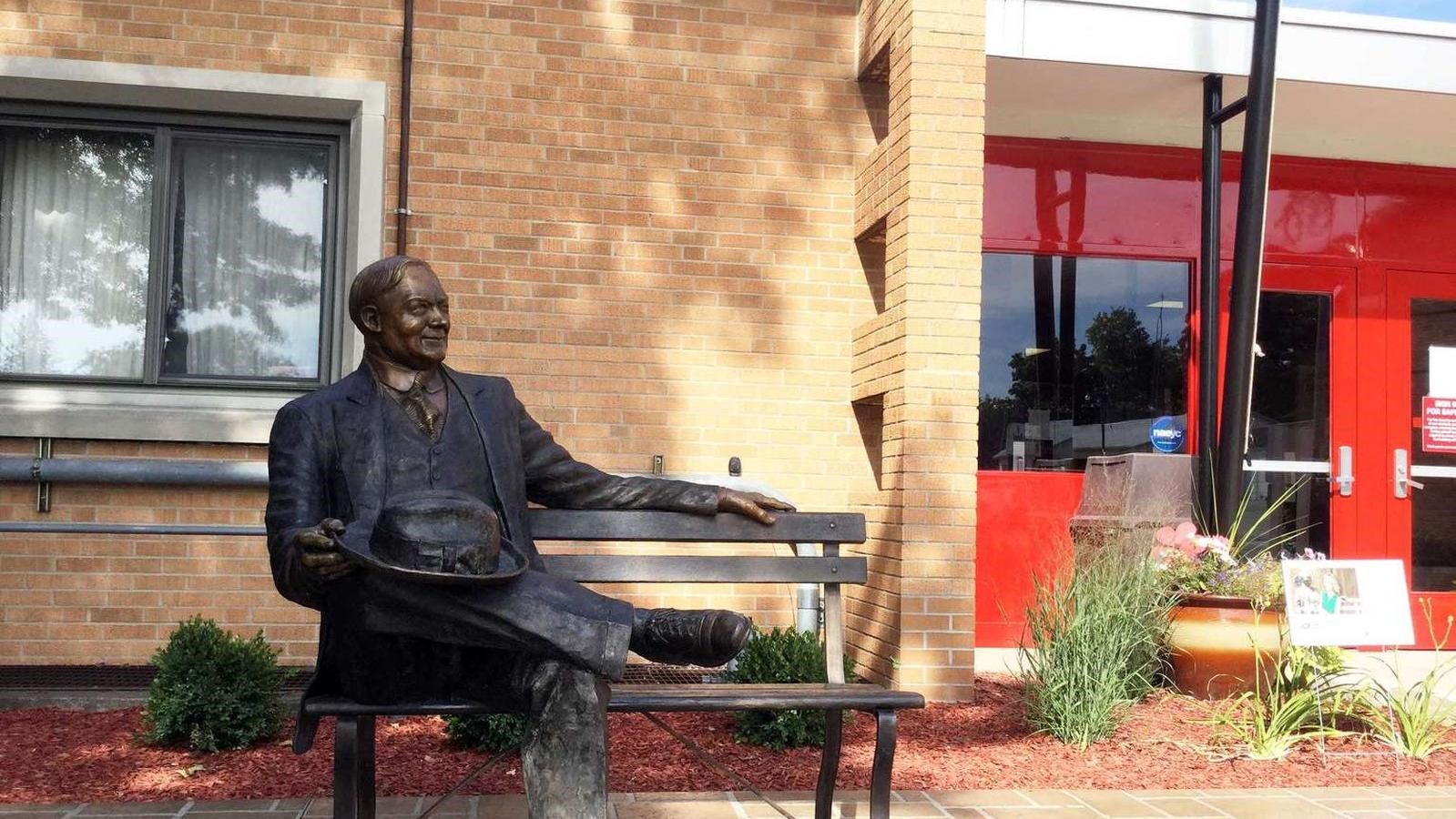 A bronze sculpture depicts a man in a suit seated on bench with room for visitors to sit next to it.