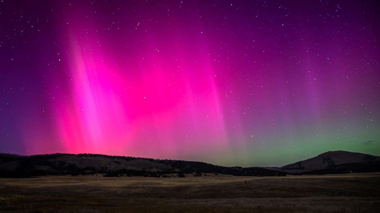 Neon ribbons of pink, purple, and green lights, the aurora borealis, loom over a valley at night.