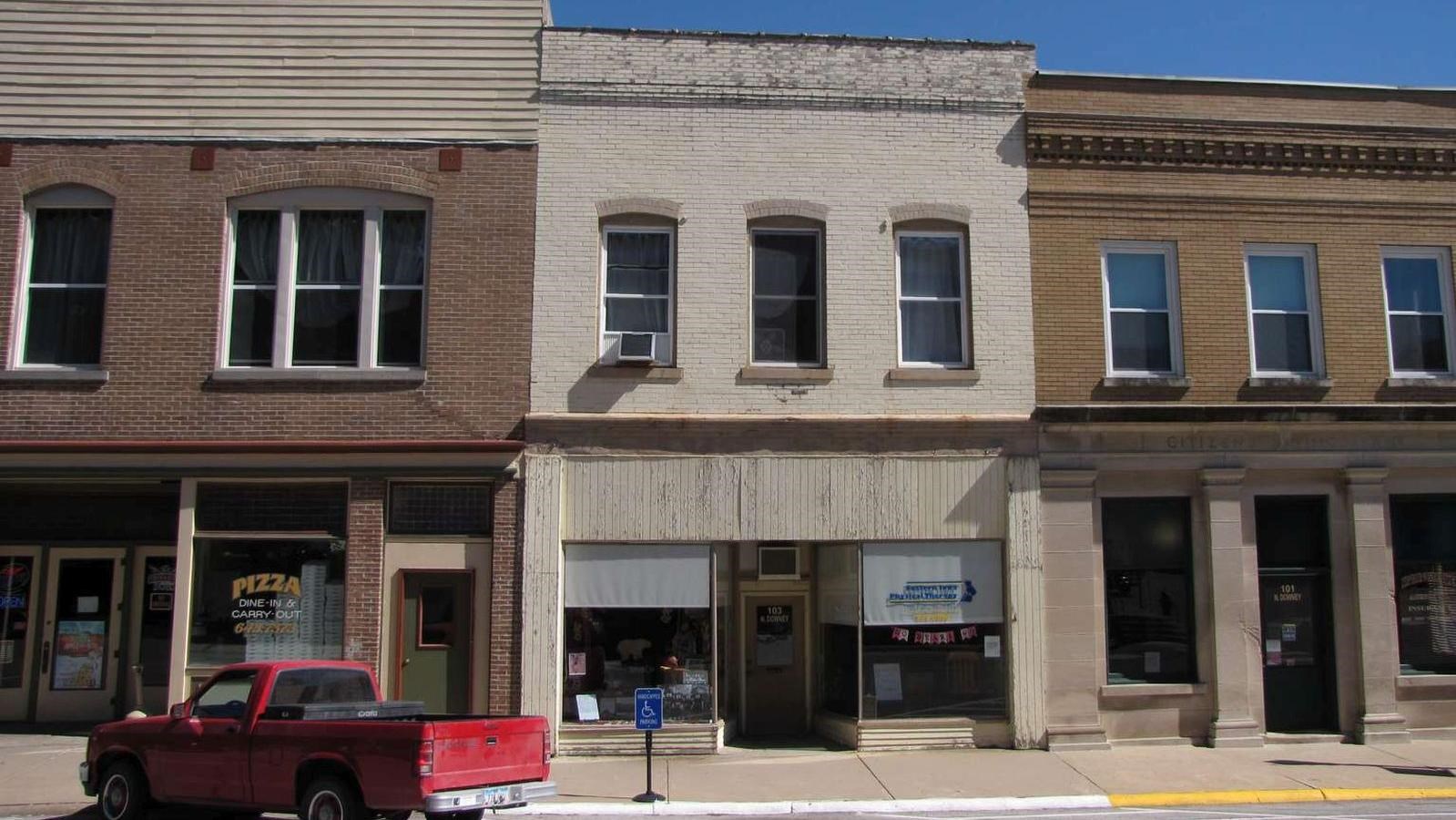 An off-white two story downtown building has a brick face over a wood-faced storefront.