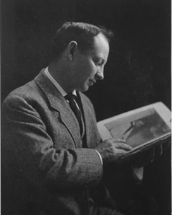 Man in dark suits poses for portrait, holding up book and reading it. 