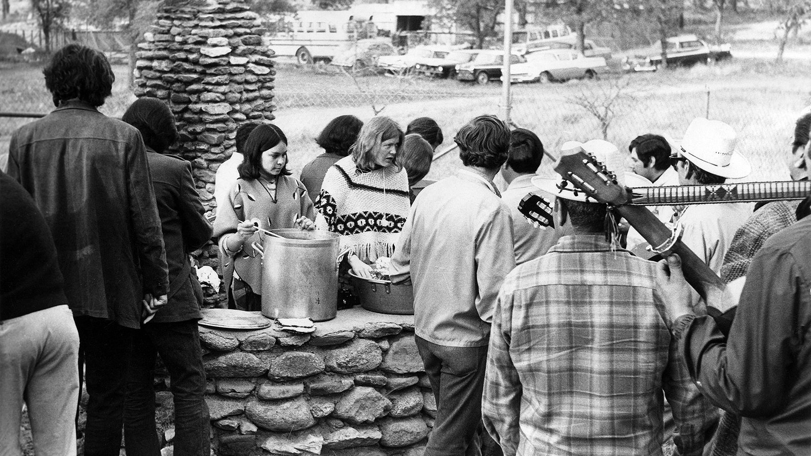 A group of people stand outdoors, waiting in line for food being served at a stone hearth