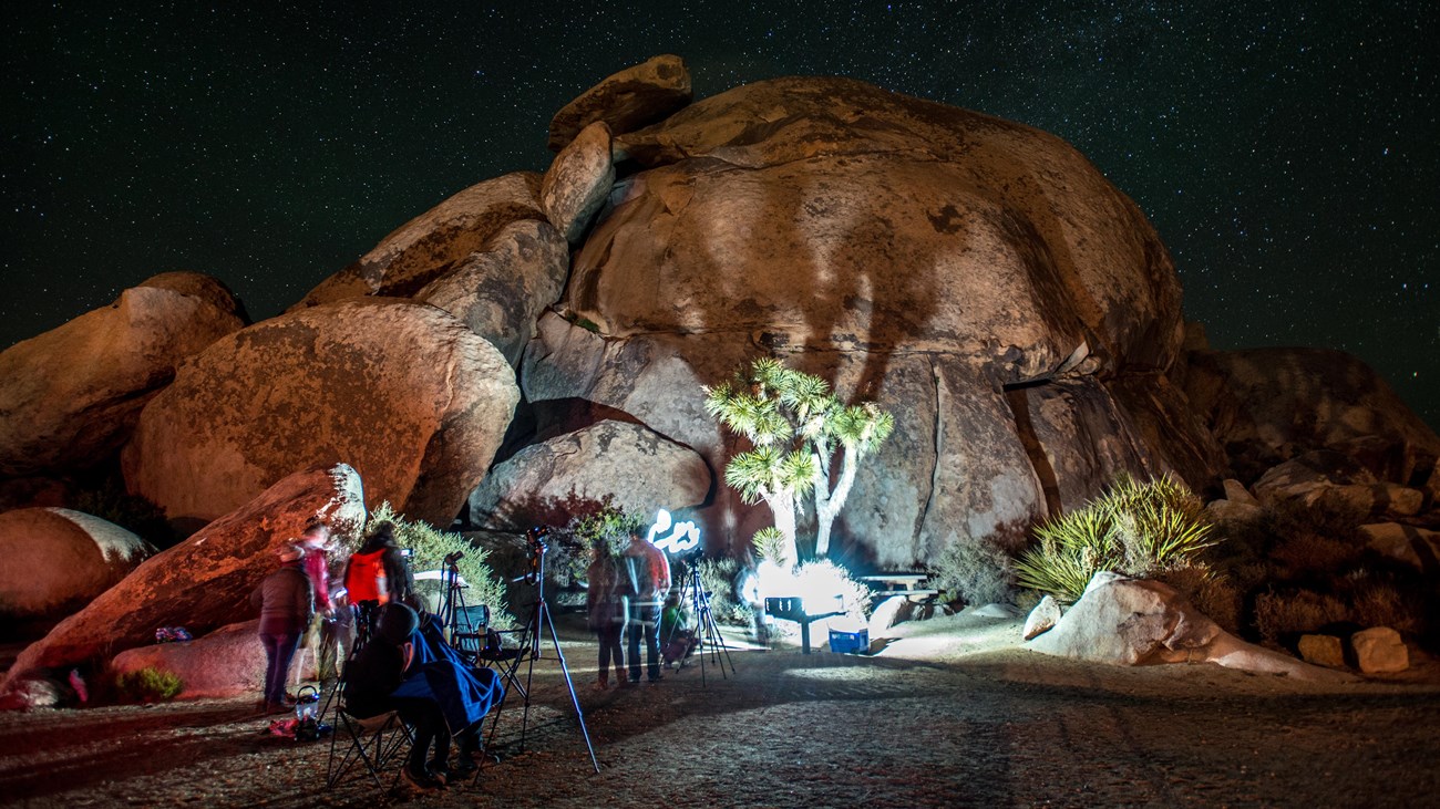 People photographing the night sky over a large rock formation and Joshua trees.