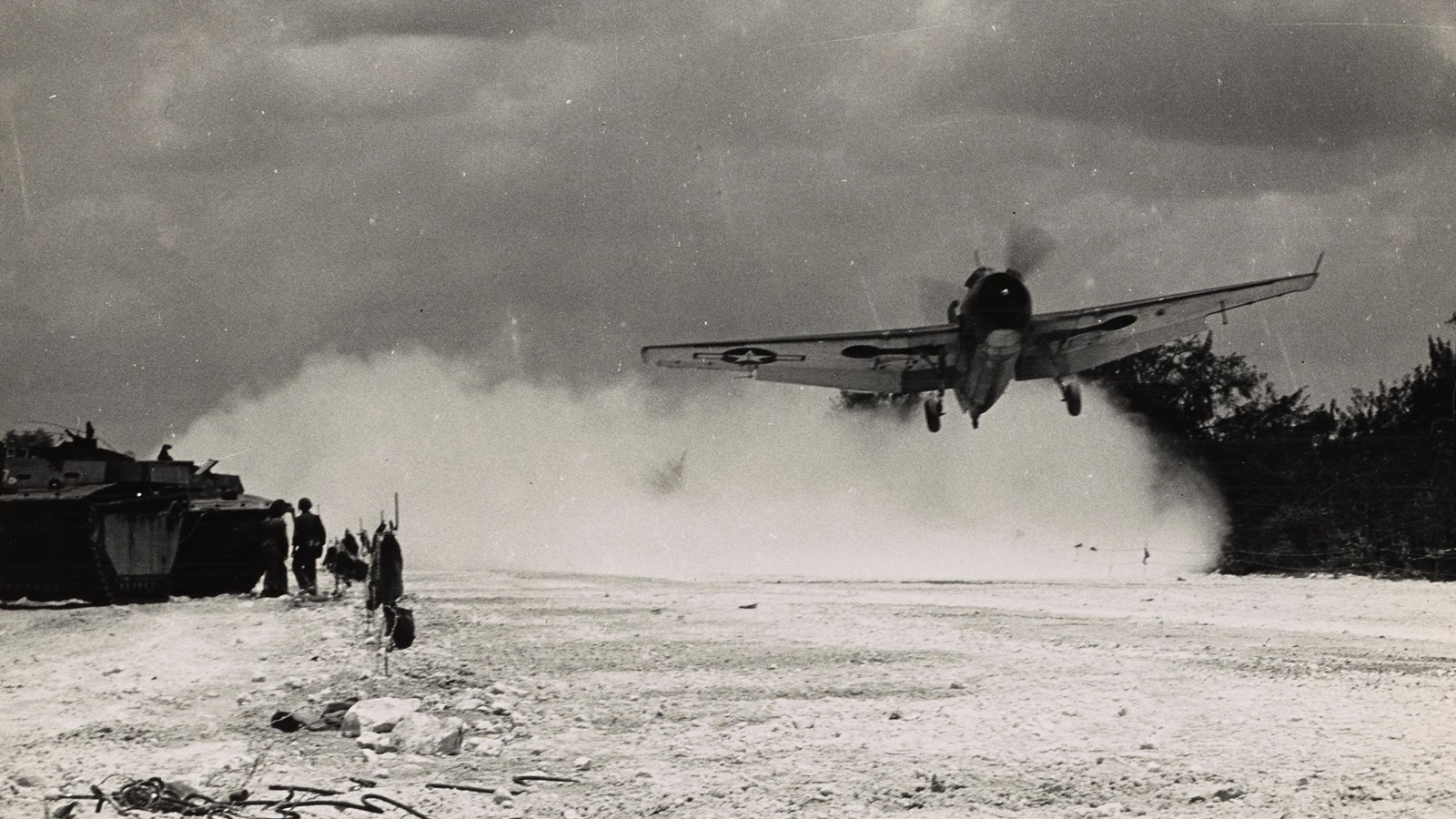 Propeller plane takes off from airfield. To the left is a tank with a US solider standing beside it.