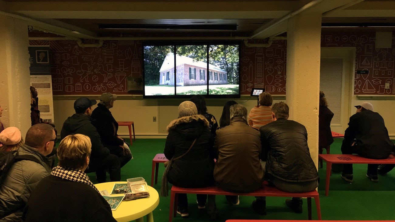 People sitting on benches watching a movie on 3-paneled television screen which shows a wooden house