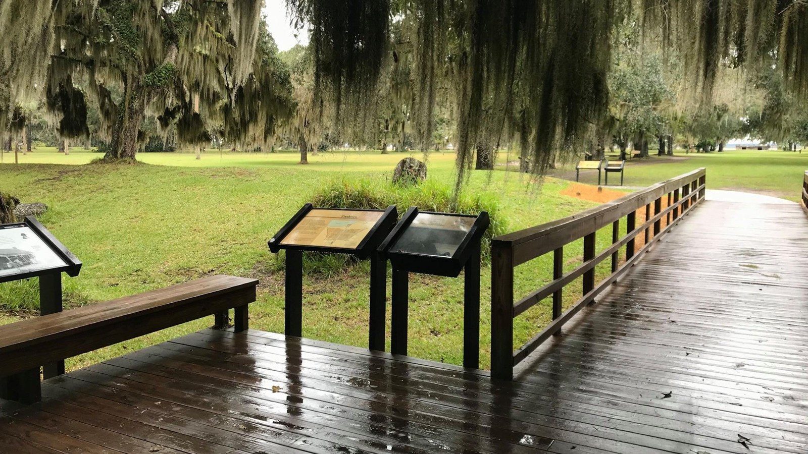 Boardwalk is lined with wooden handrails, displays, and bench; grass and trees surround.