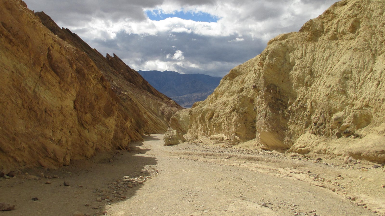 A gravel wash winds between bare yellow hills, with dark clouds overhead.