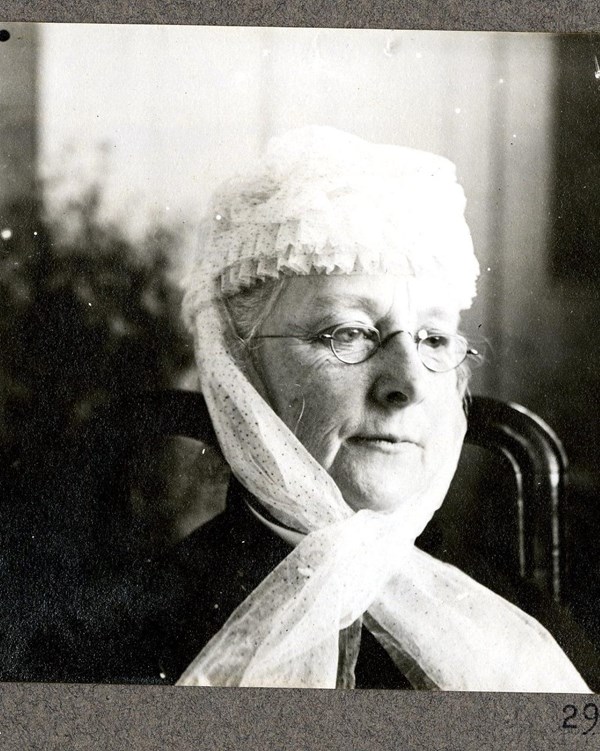 Elderly woman wearing glasses and a white scarf draped over her head poses for a picture.