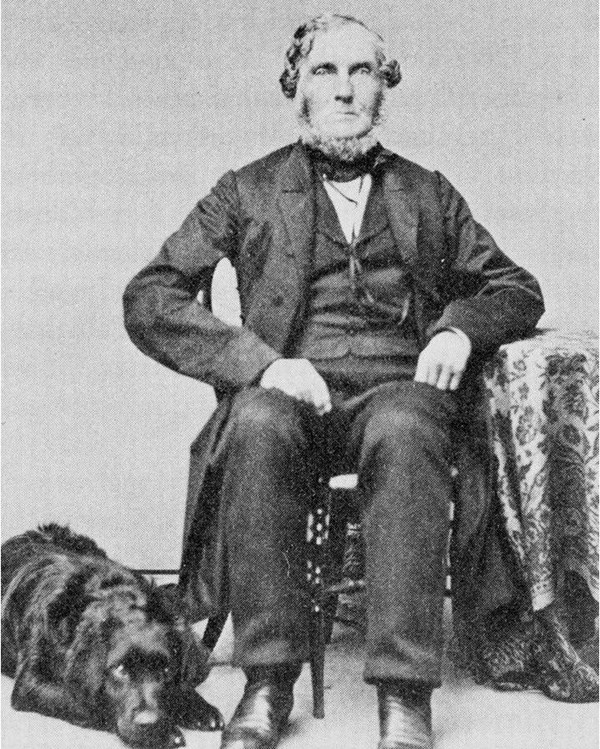 Elderly man in suit with short white hair sits at chair with black dog laying by feet.
