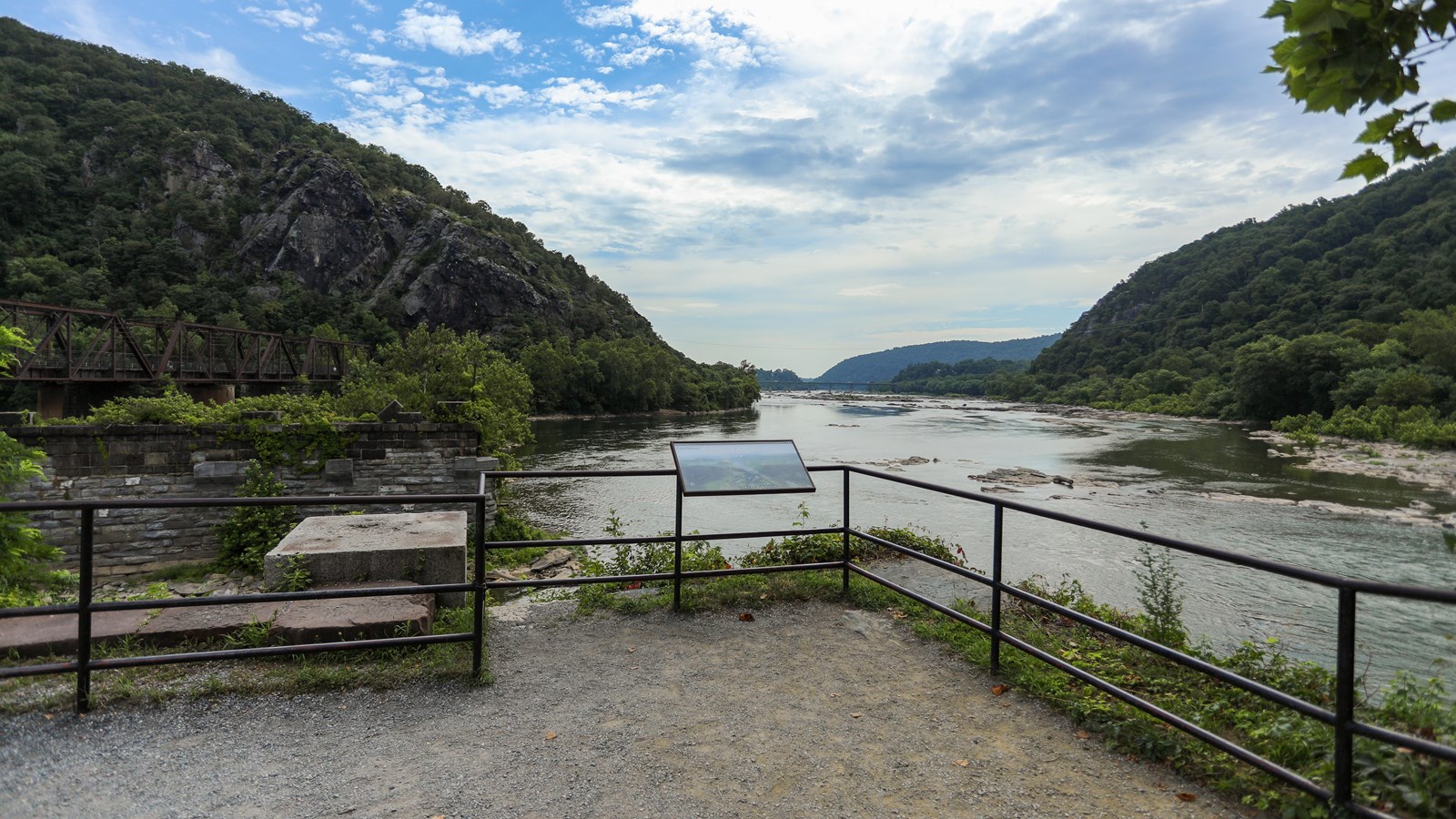 An area with loose gravel and a metal railing overlooking a wide river valley lined with trees.