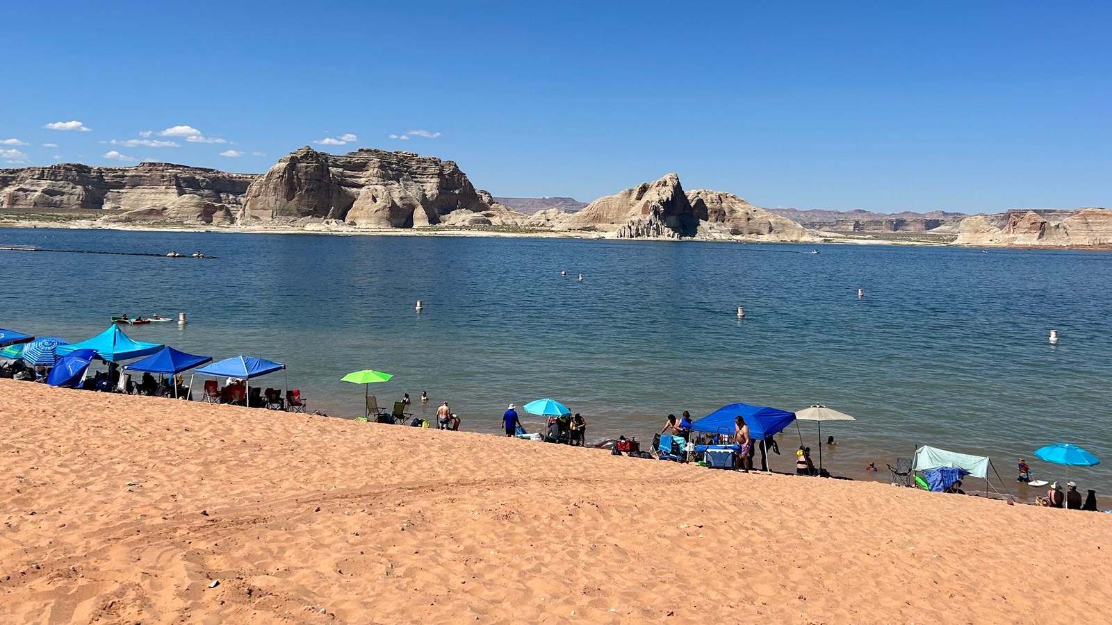 A sandy beach on a desert lake. People and umbrellas on beach. Sandstone buttes in background.