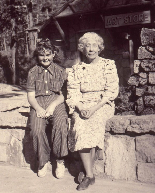 A young girl and an older woman sitting on a rock wall backed by a building with a sign: "Art Store"