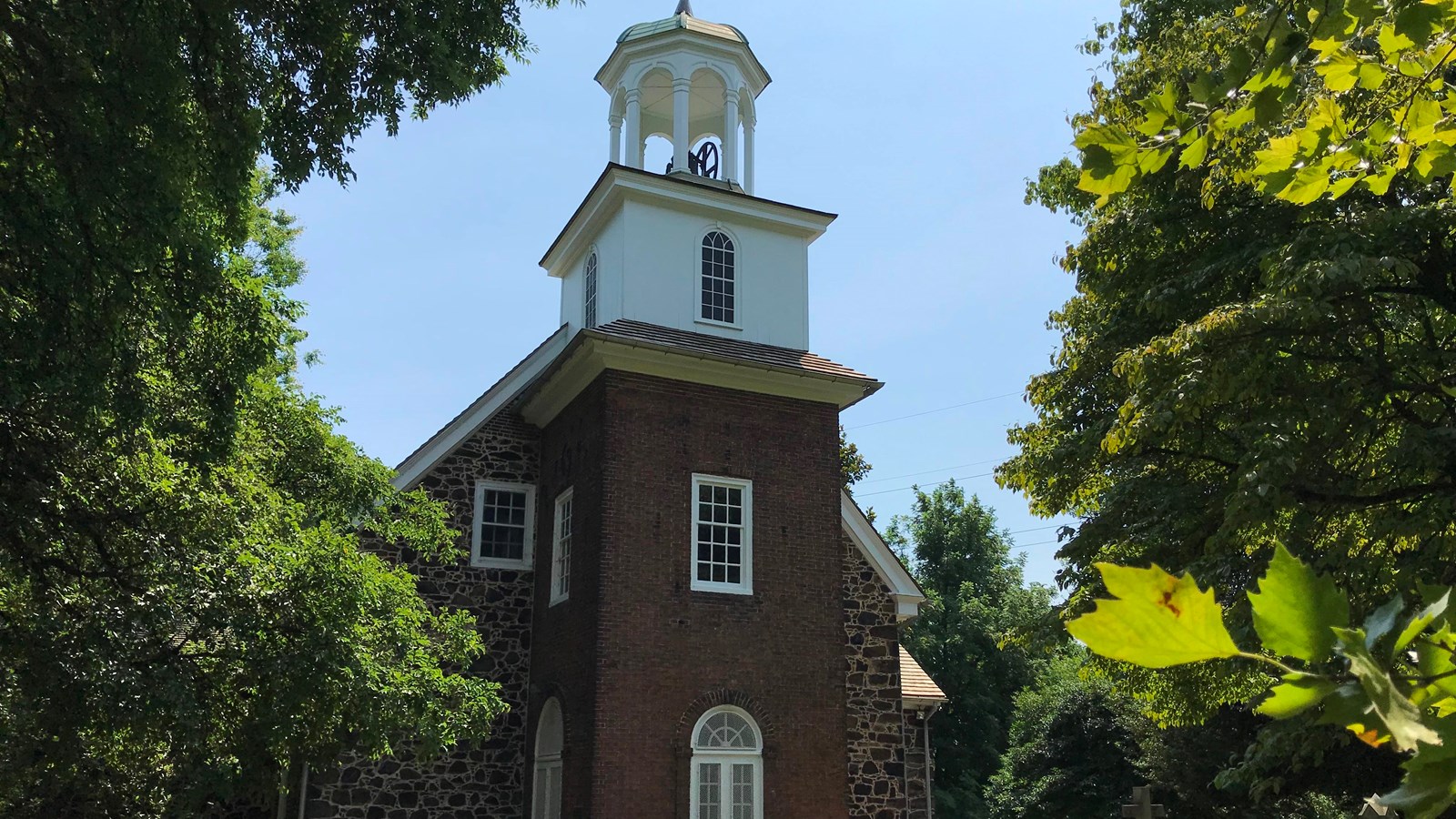 A brick church with a white cupola is outlined by green leaves. 