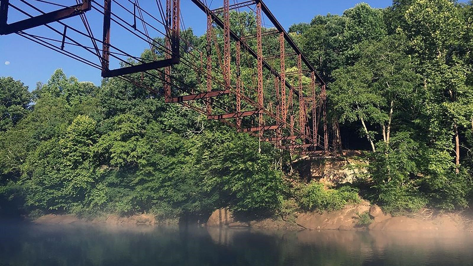 View looking up at steel skeleton of bridge over foggy river with wooded shore in the distance.