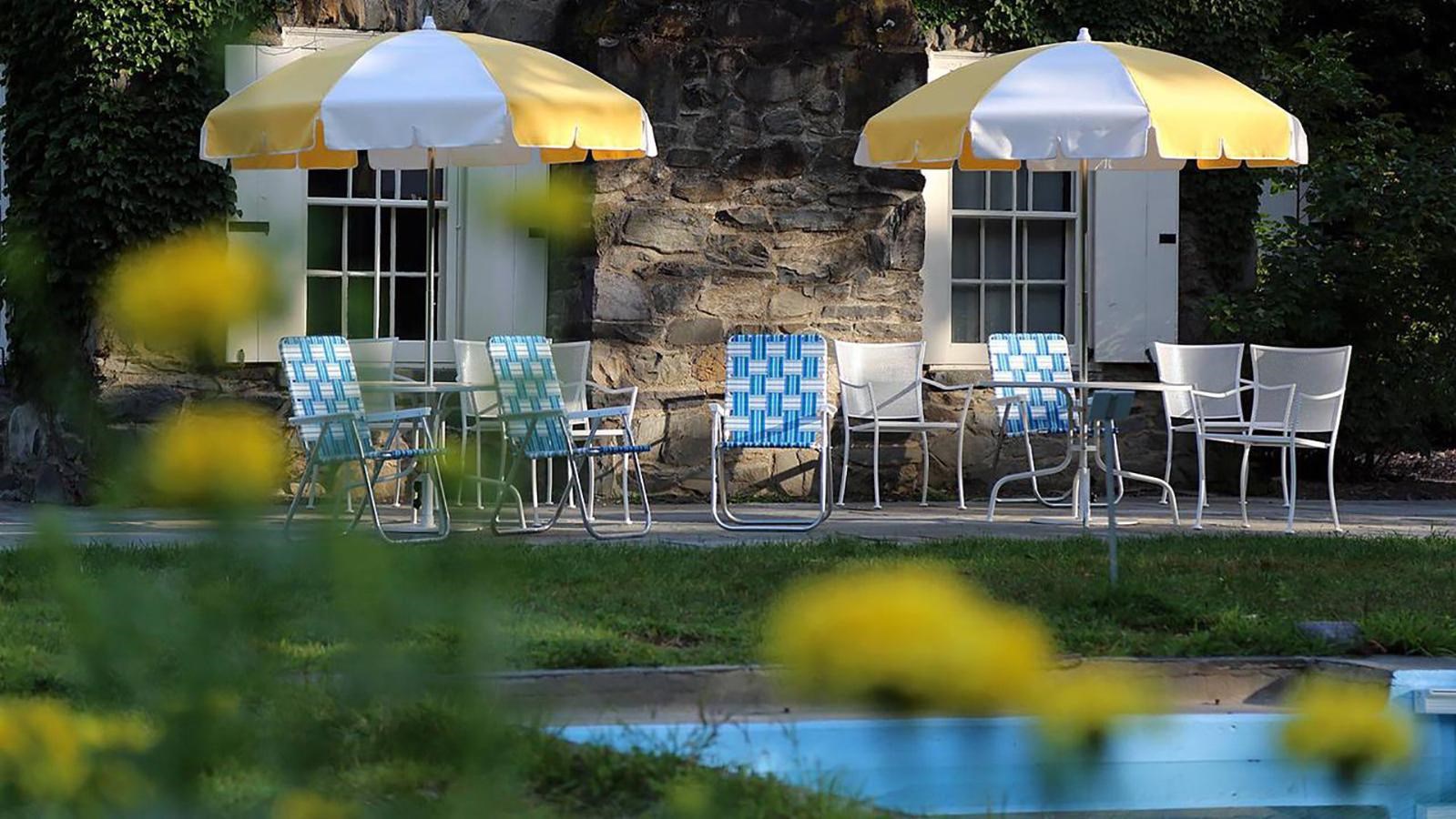 A group of tables with umbrellas and folding chairs on the edge of a swimming pool amid a grass lawn