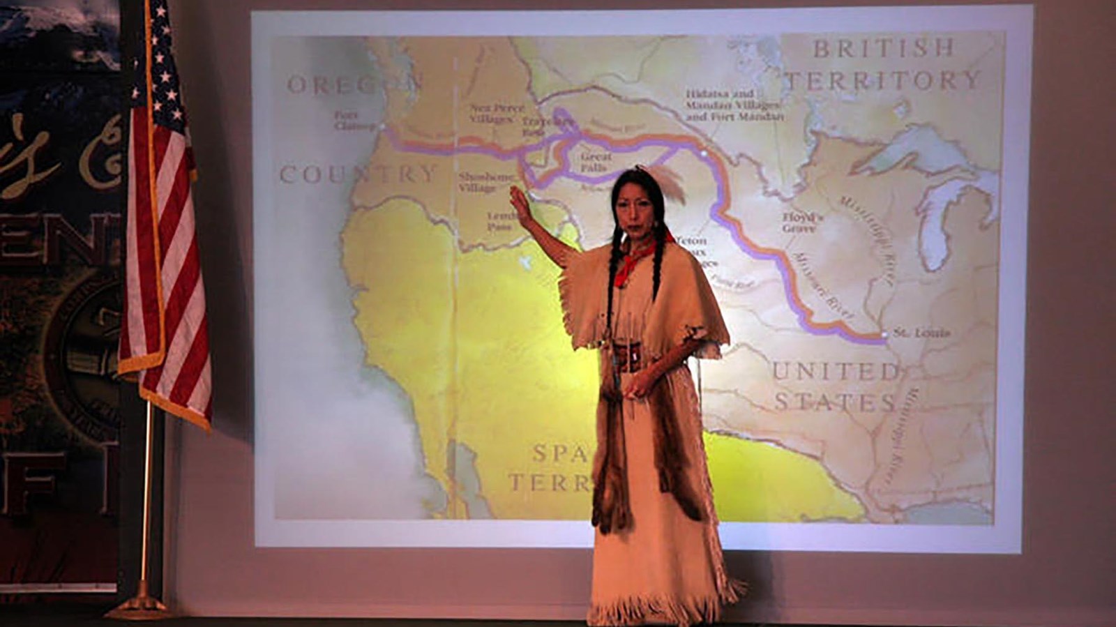 Woman in Native American attire stands on stage in front of screen pointing at a location on a map. 